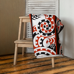 New Zealand Beach towel with a red, black, and white pattern draped over a wooden stool on a wooden floor.