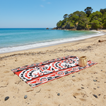 Maori Beach towel with red and black Kōwhaiwhai patterns on a sandy beach with ocean and trees in the background