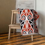 Maori Beach Towel with red, black, and white patterns draped over a wooden chair on a wooden floor.