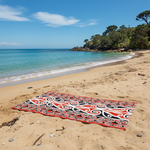 Maori Beach towel with red, black, and white pattern on a sandy beach with ocean and trees in the background