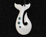 Front view of a hand-carved white bone pendant combining a whale tail and fish hook with three round iridescent paua shell inlays on a black koru patterned background.