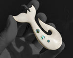 Three-quarter angle close-up of a carved bone whale tail fish hook pendant held in a gloved hand, highlighting the smooth curved finish and green-blue paua shell insets.