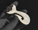 Reverse side of the hand-carved bone whale tail fish hook pendant held in a hand, showing the smooth, unadorned back surface and top suspension cord hole.